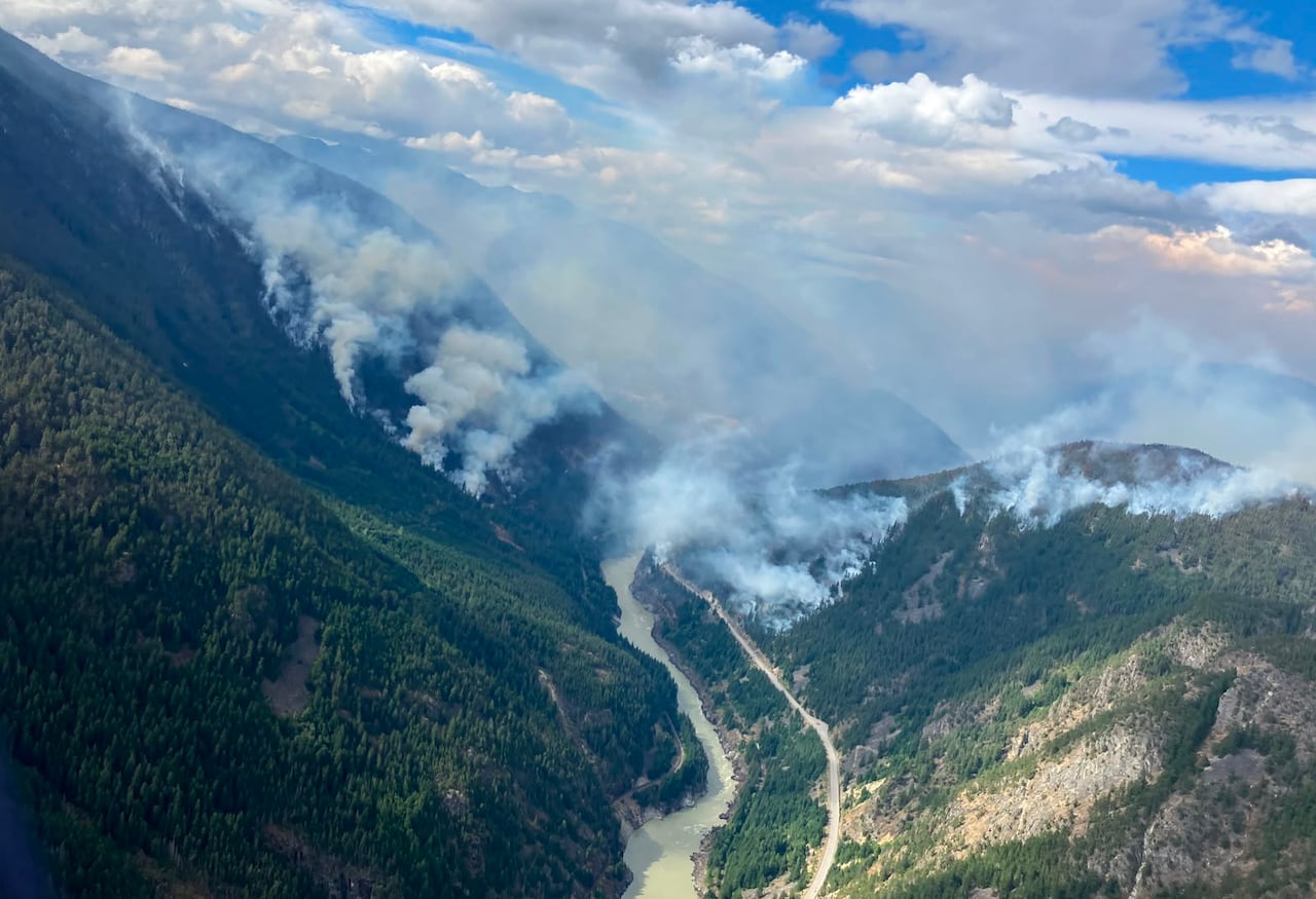 Wildfire smoke rise from treed mountainsides, with a river running down a valley.