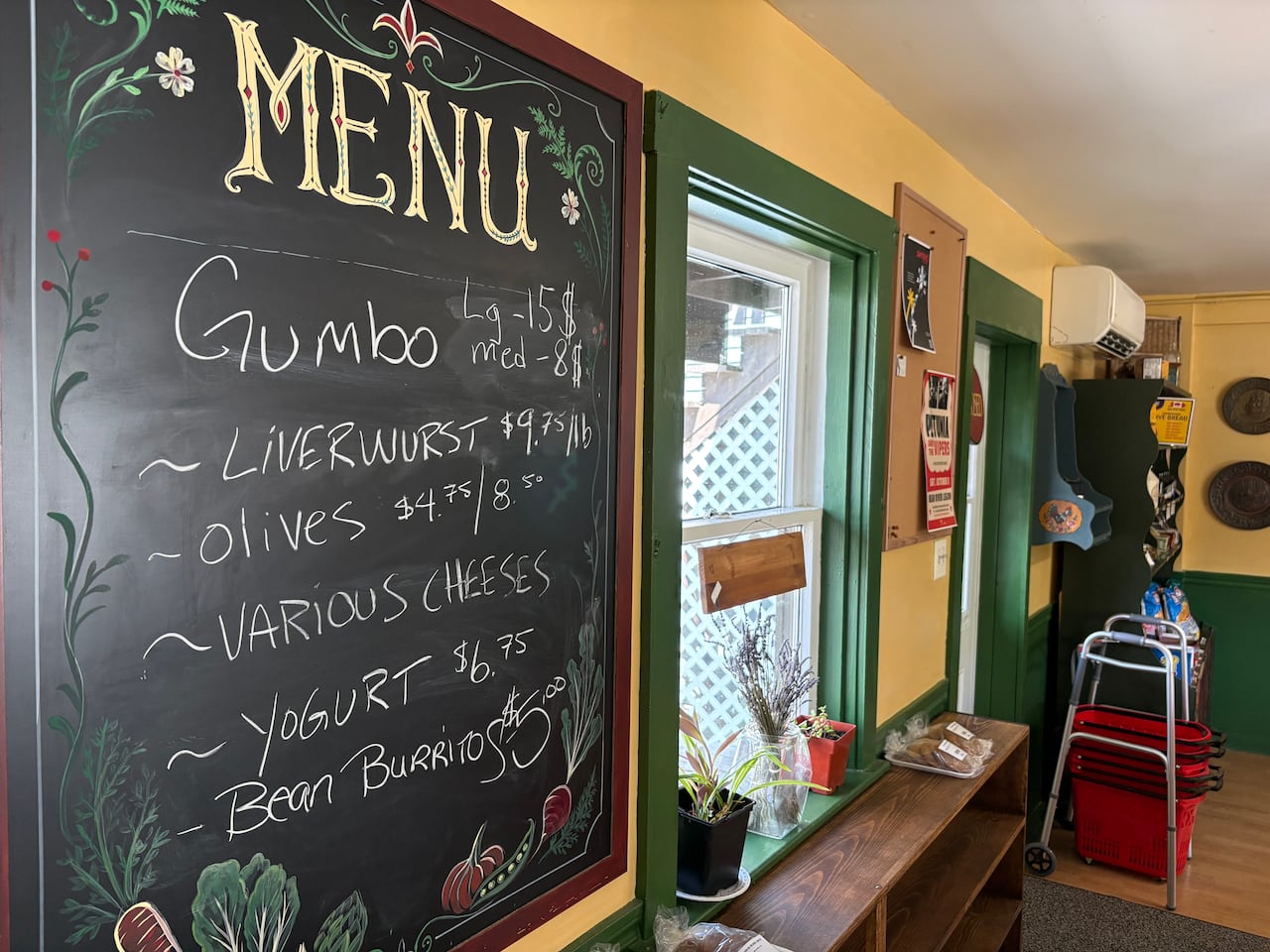 A menu sign notes prices for gumbo, liverwurst, olives, cheeses, yogurt and bean burritos.