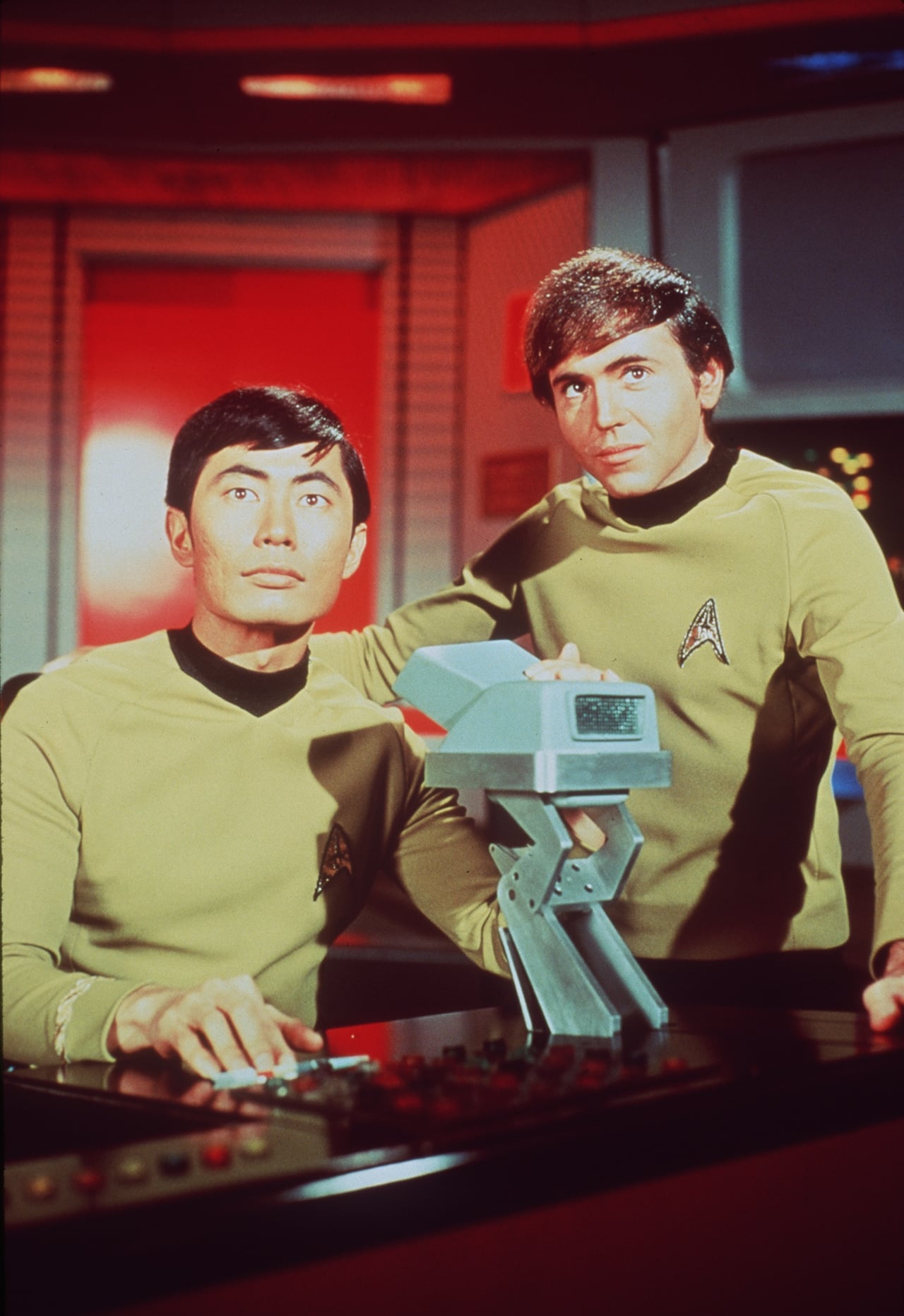 An Asian man and a European man in green uniforms on the set of a sci-fi show.