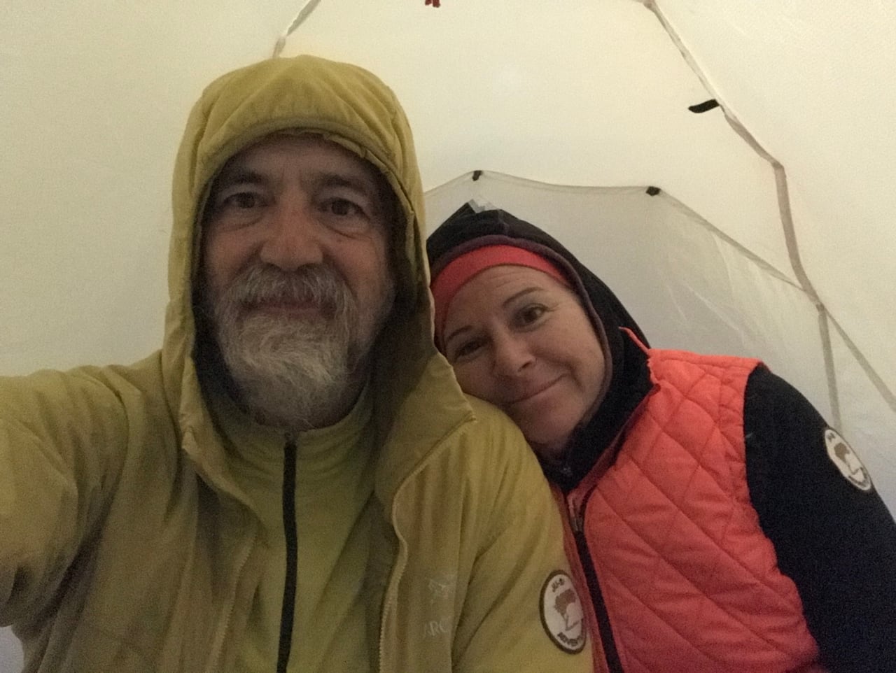 Two hikers in rain jackets take a selfie while resting inside a tent