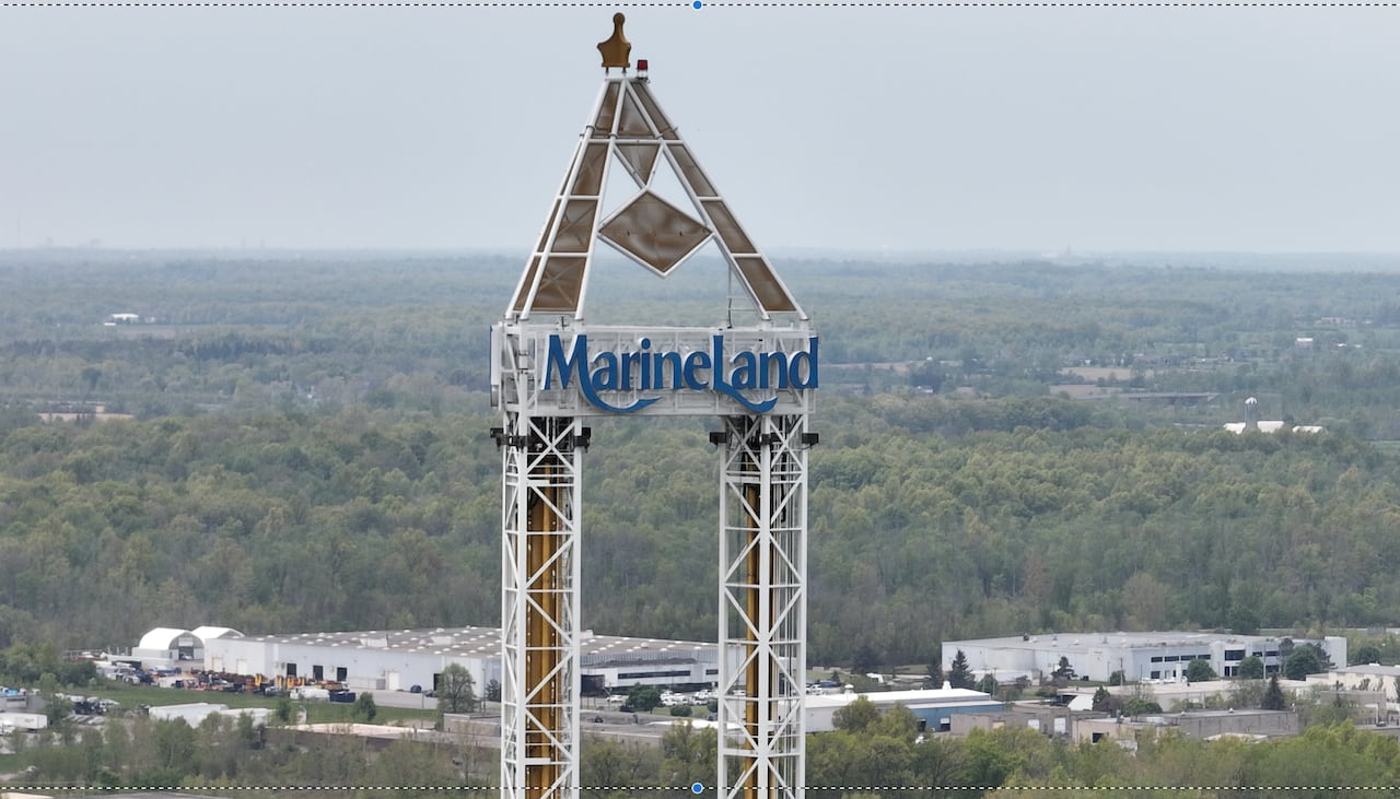 An amusement park drop tower ride features the Marineland logo on its white structural top, set against a background of green trees and industrial buildings.