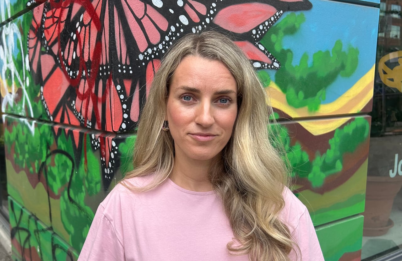 A woman with long blond hair wearing a pink top stands in front of a wall painted with a colourful mural of a butterfly.