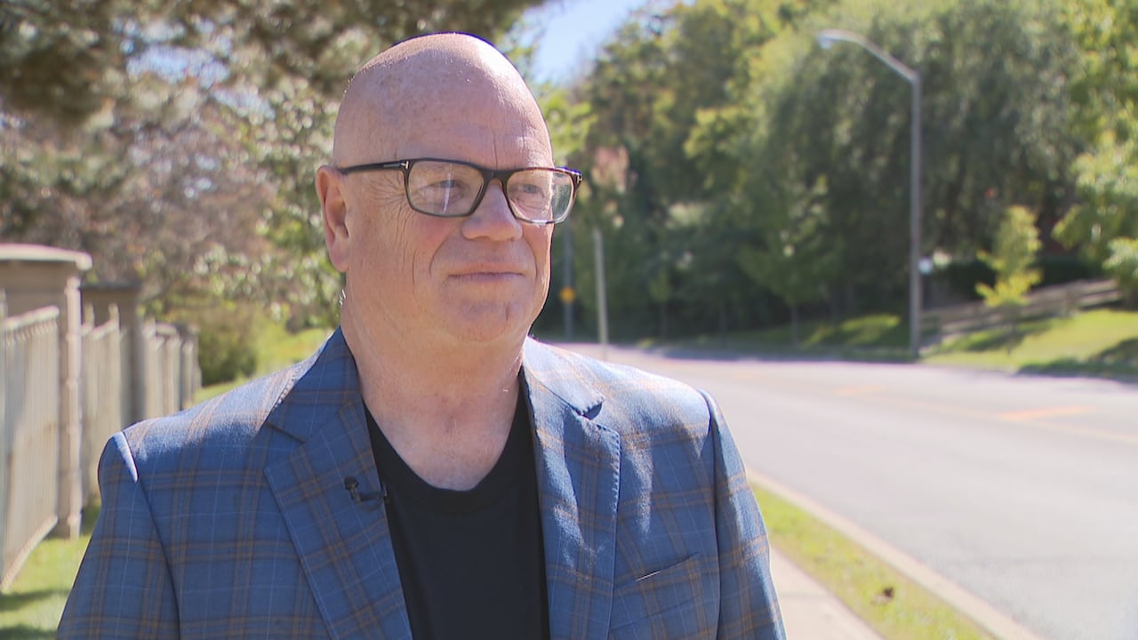 A man wearing glasses standing next to a residential street with trees in the background.