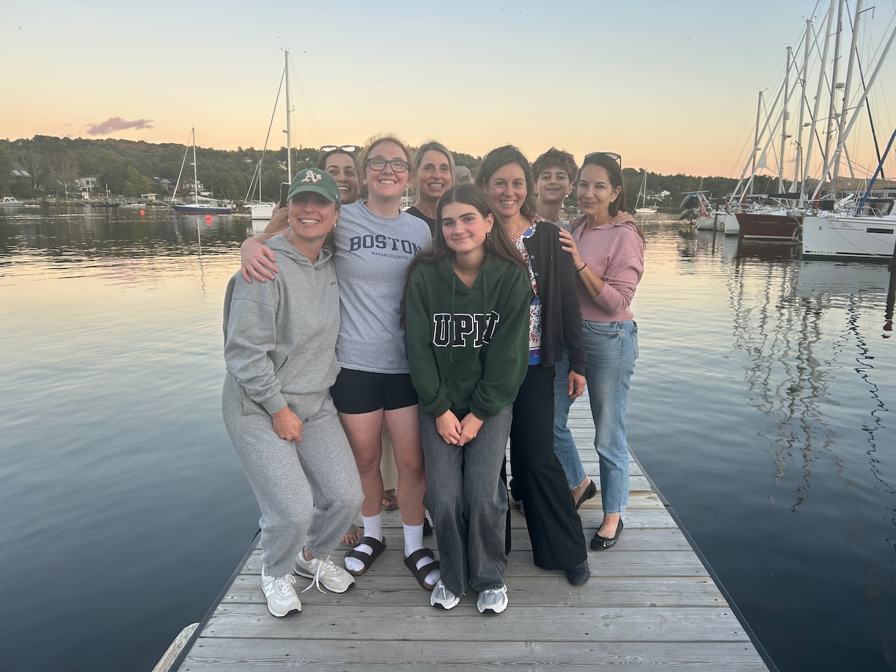 A group of people pose for a photo on a wharf. Behind them is the water and sailboats.