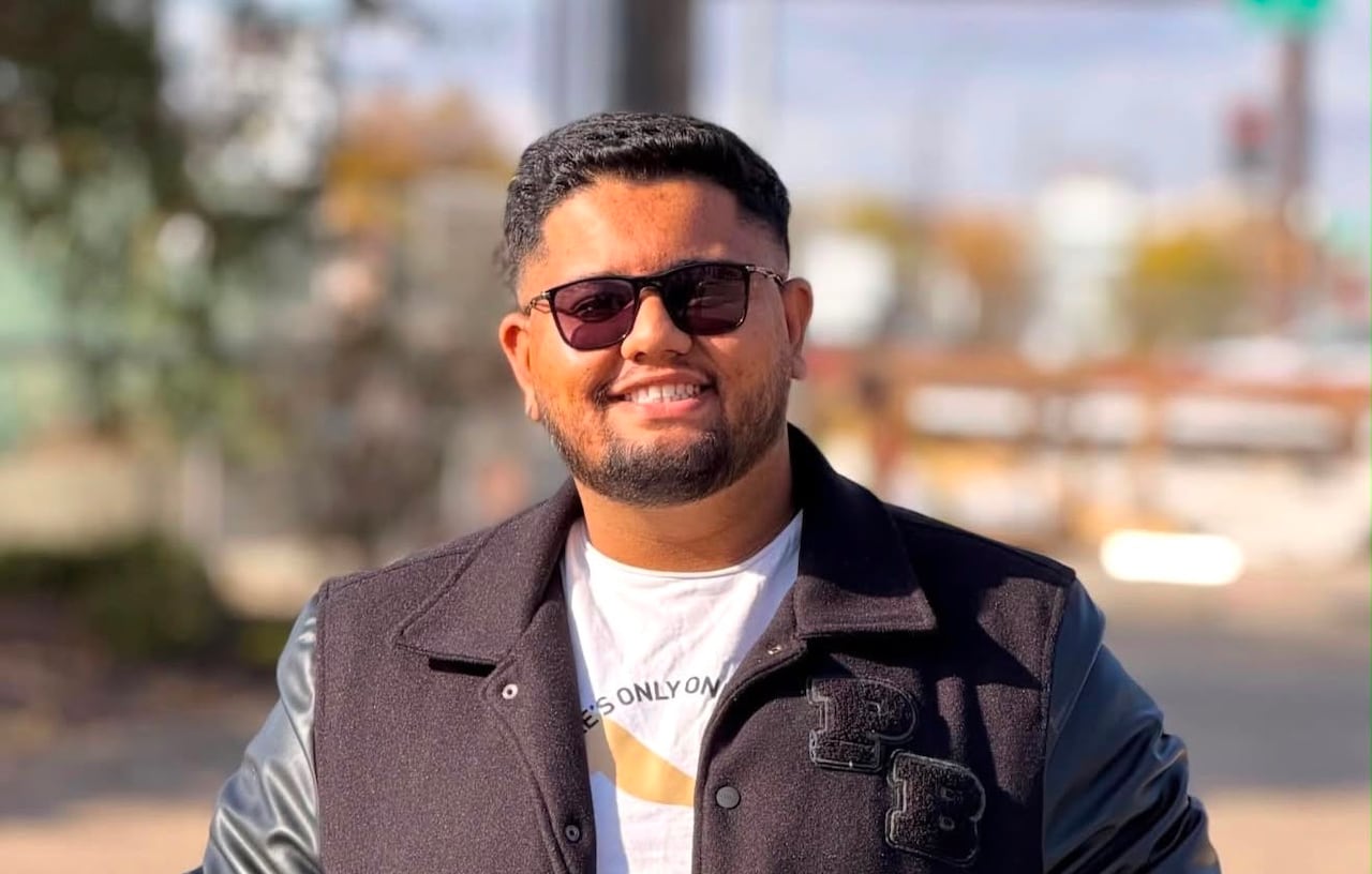 A man wearing sunglasses with his hands in his pockets stands smiling.