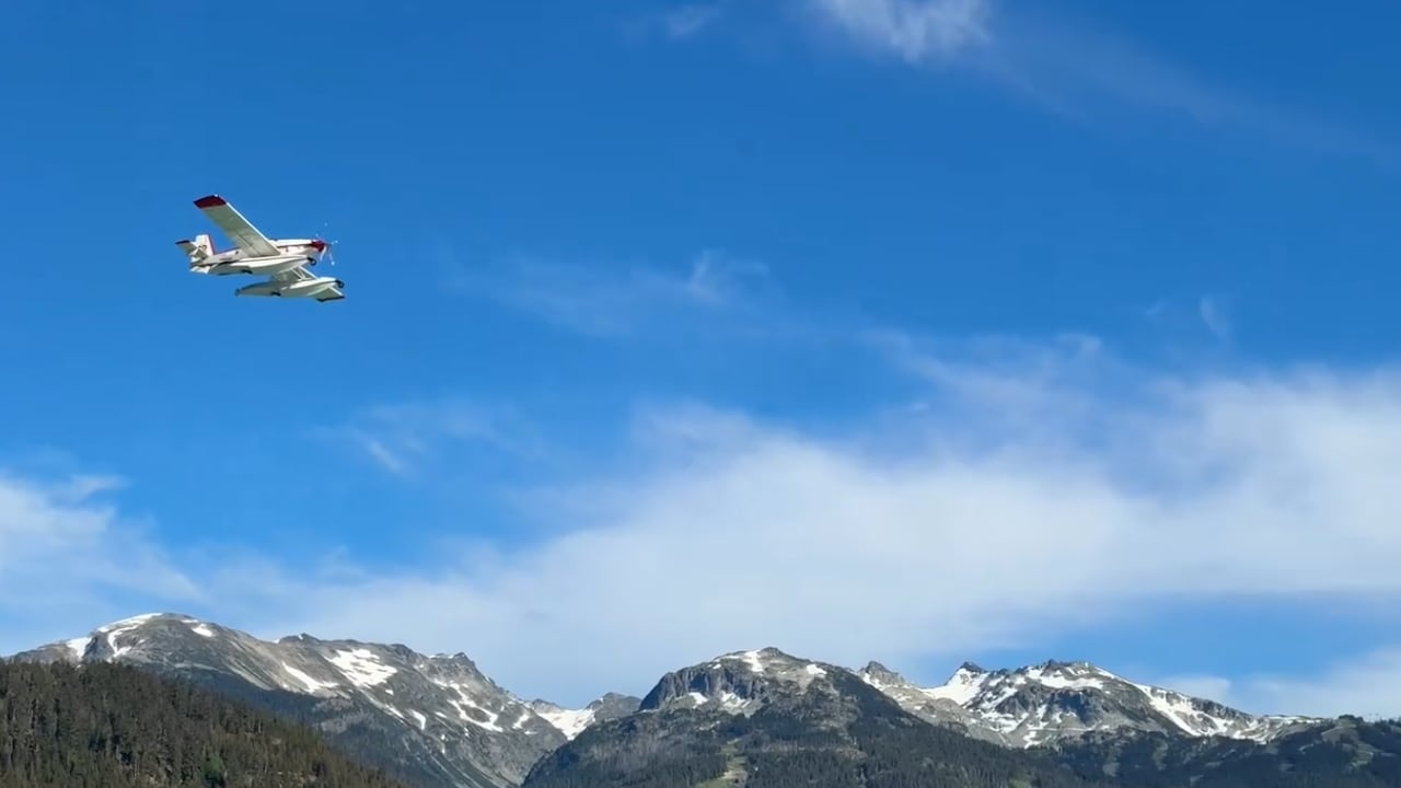 A small white plane with pontoons flies with mountains in the background.