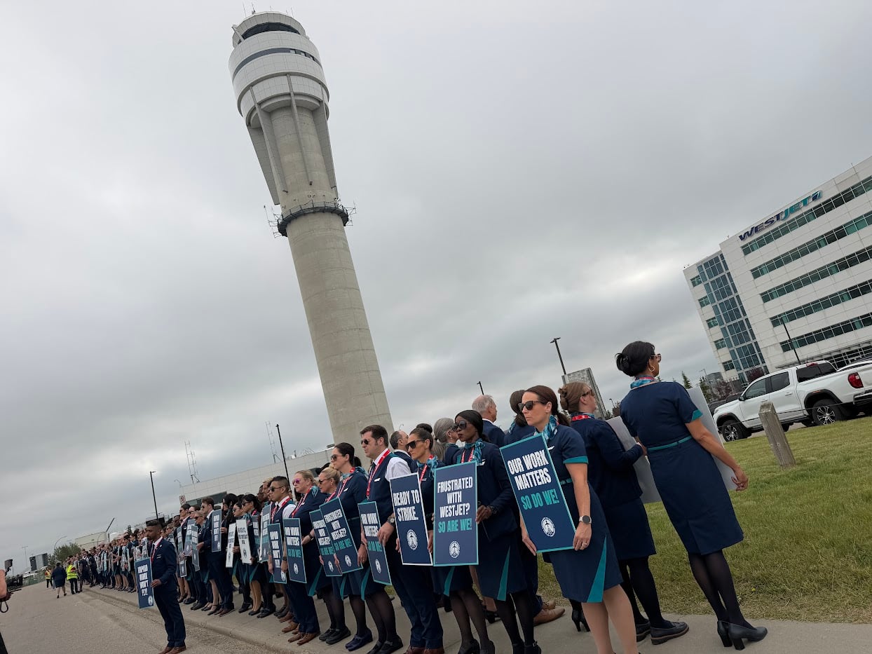 Flight attendants line up outside wearing union picket signs saying things like "ready to strike." The WestJet office is pictured in the background.