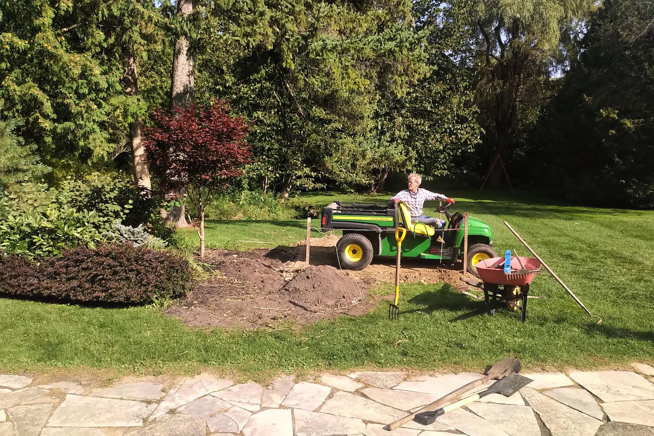 A man on a small green tractor in a patch of dirt near trees and grass
