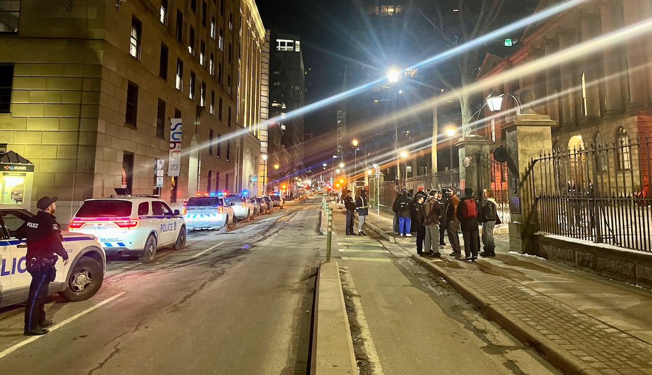 People stand outside on a sidewalk as police cars are parked across the street.