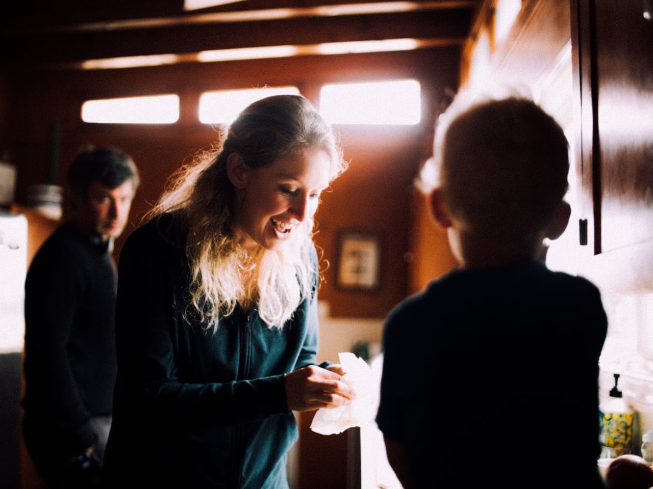A smiling woman can be seen interacting with the silhouette of a child. A man stands behind her, watching them.