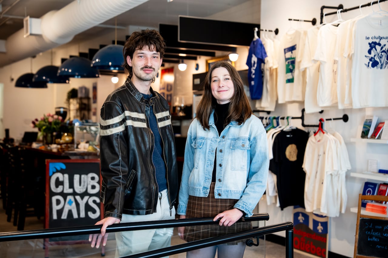 A man and a woman stand in a room with t-shirts hanging from the wall, and a sign that says Club Pays behind them