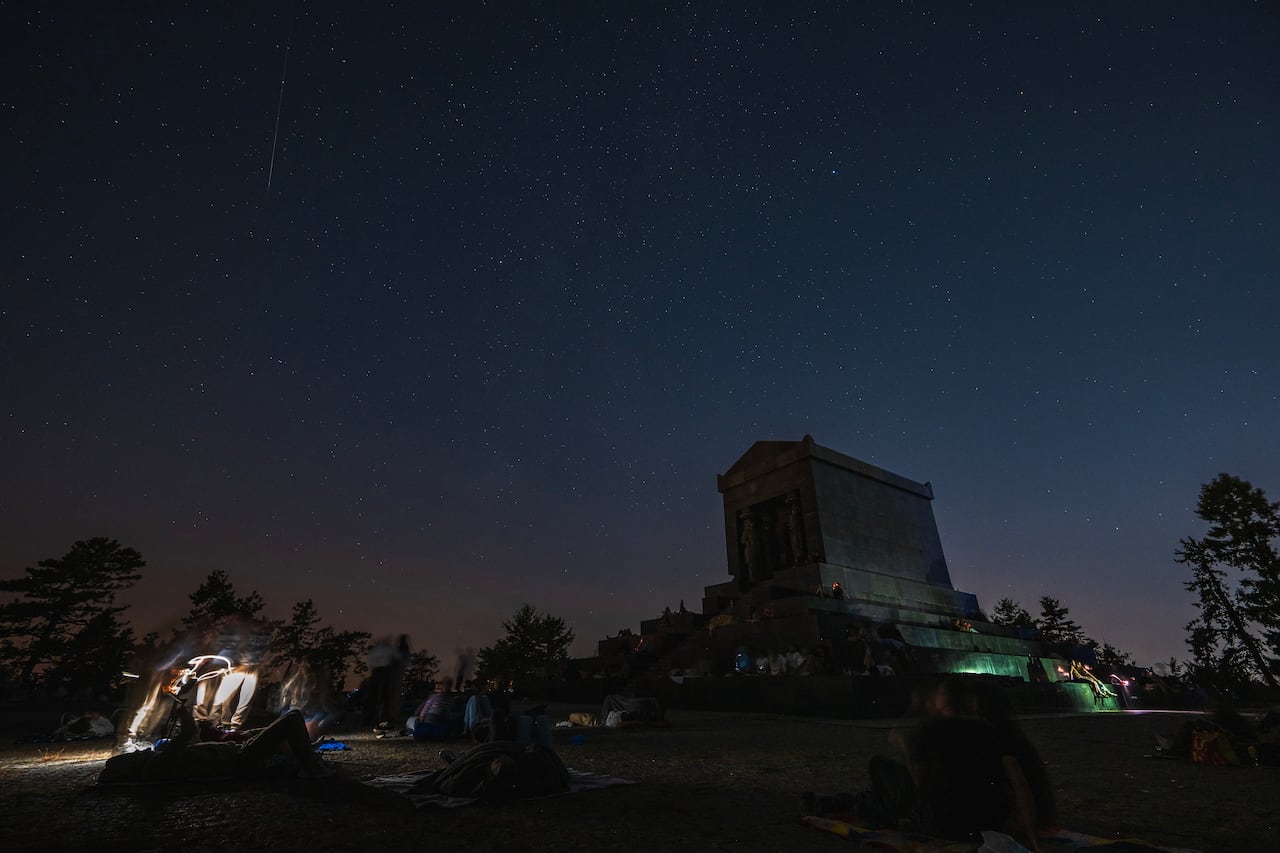 Meteor streaks and stars at night, with people gathered on a grassy area by a monument.