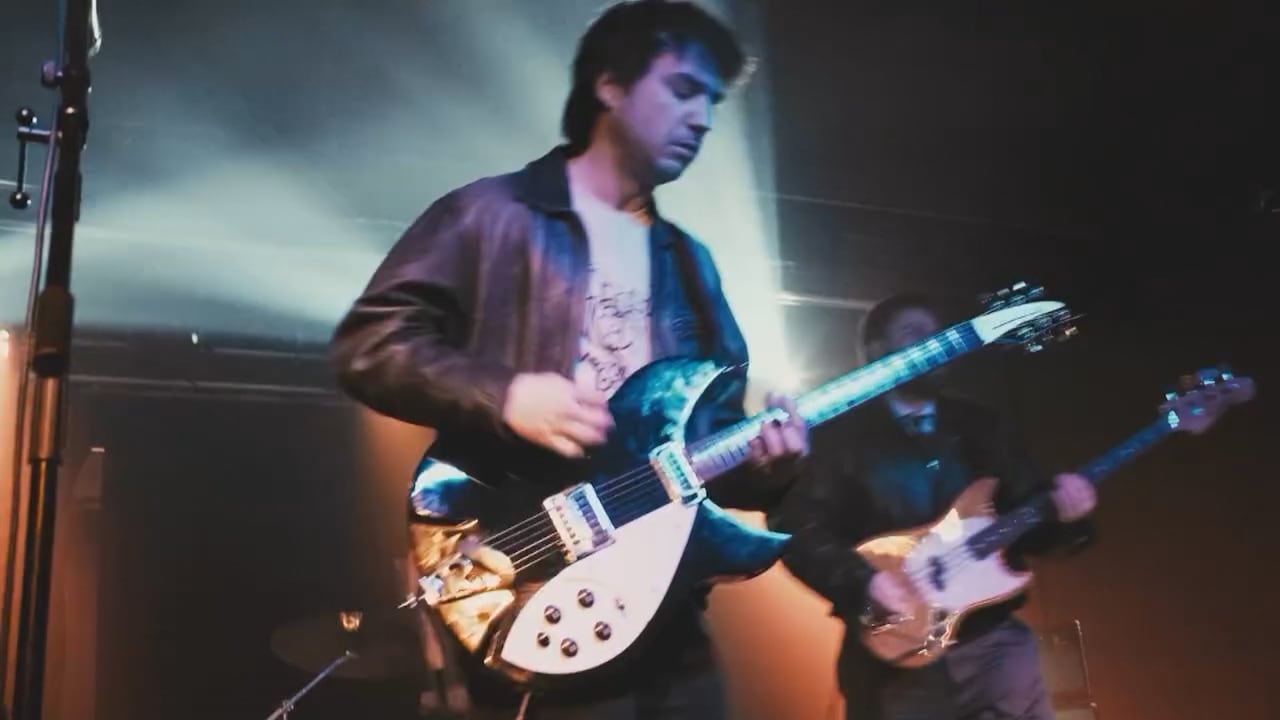 A man in a black jacket plays guitar in front of  another man in a black jacket playing bass guitar with bright lights in the background.