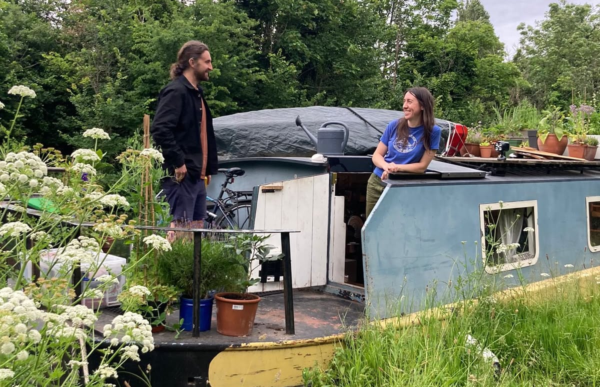 A man and woman are pictured on a houseboat amid lush greenery.