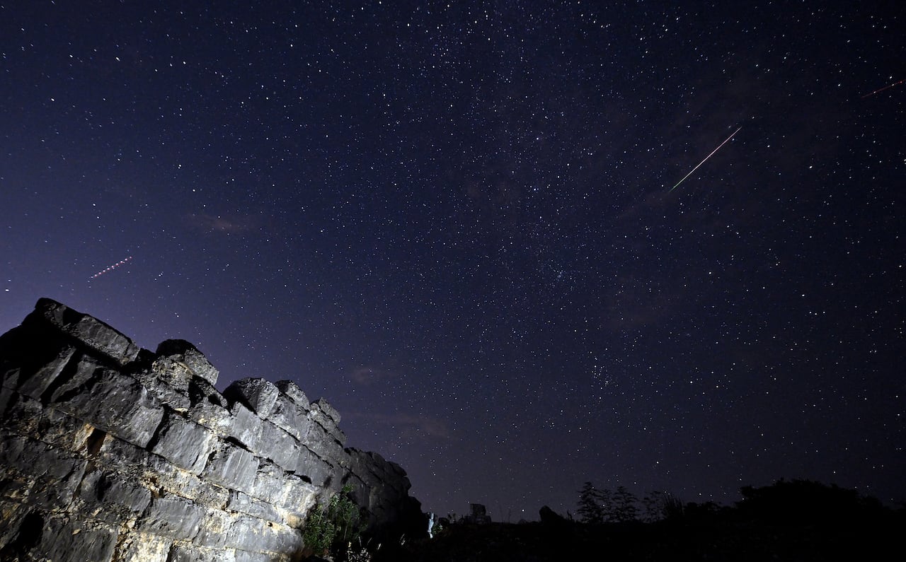 Meteor streaks and stars in the night sky above stone walls that are lit.