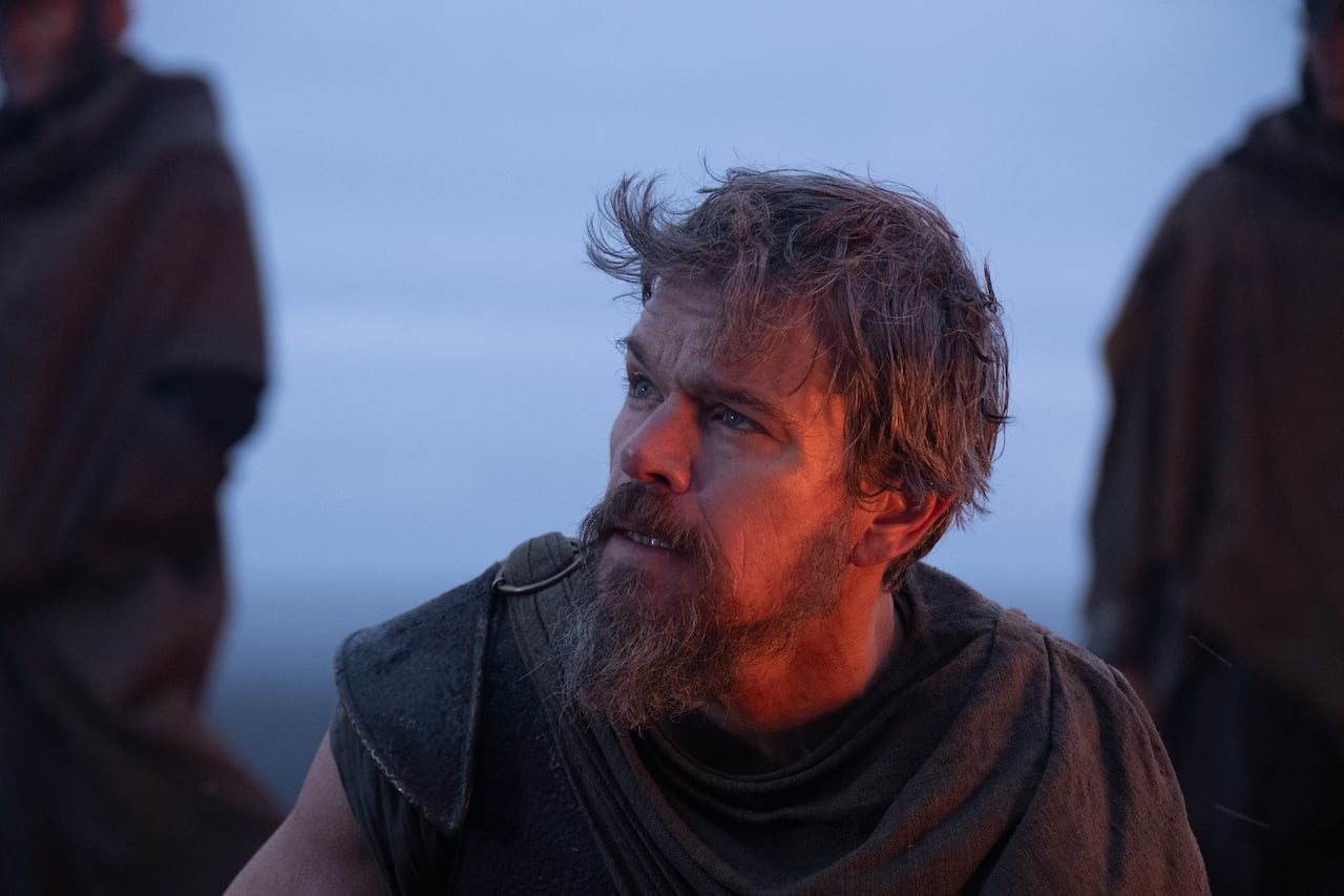 A man with a weary expression and grizzled beard,  near the ocean looks off to the distance.