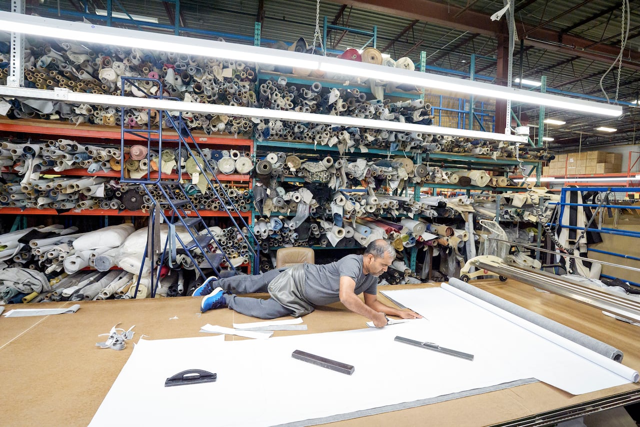 A man lying on a table cutting fabric, surrounded by rolls of fabric on shelves in a factory.