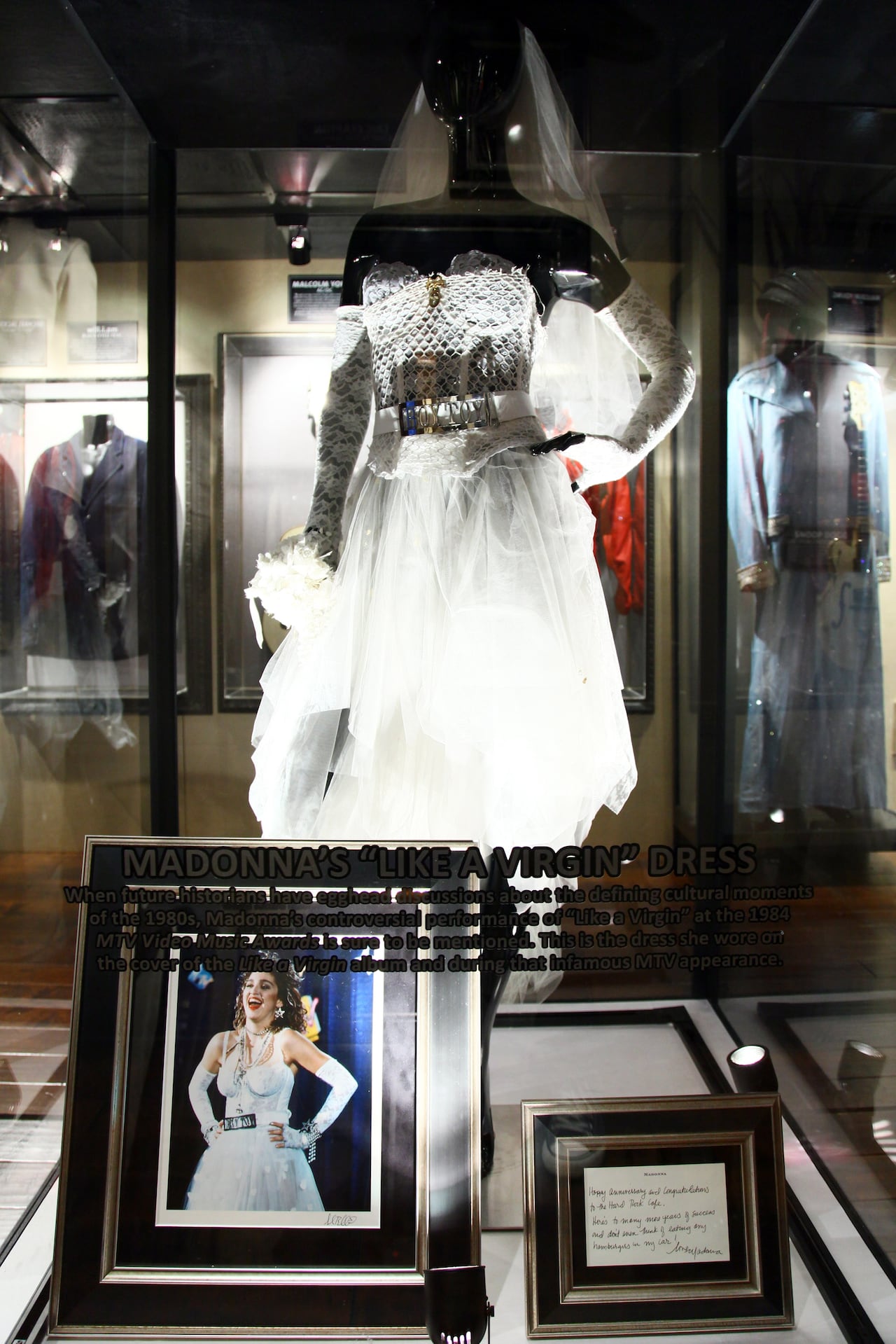 A wedding dress and a belt reading "Boy Toy" displayed in a case on a mannequin. A photo of a woman wearing the dress and a plaque sit in front of it.