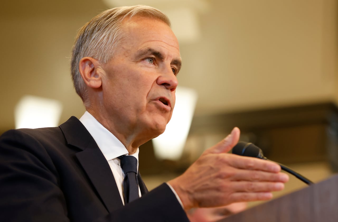Man with white and grey hair wears a black suit with a white shirt and black tie while speaking into a microphone at a podium