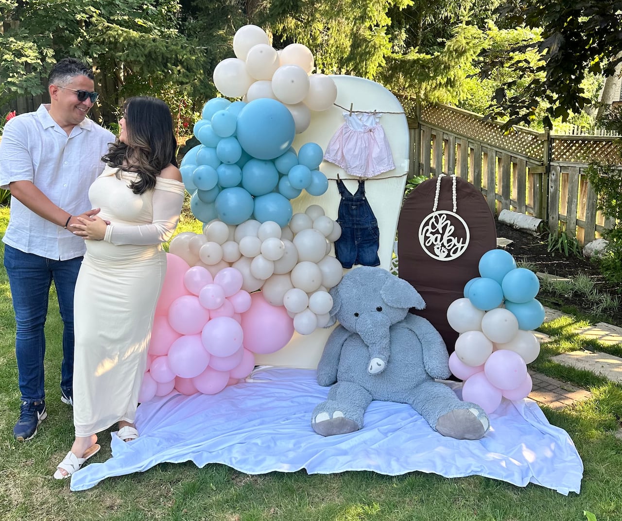 A woman and man stand in a backyard next to a baby shower display