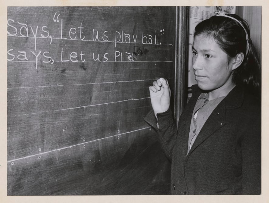 Black and white photo of a First Nations girl at a classroom chalkboard.