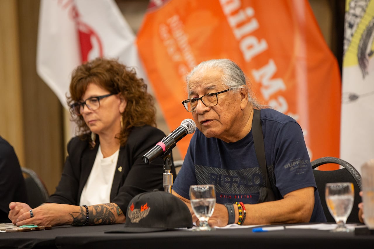 Two people are seen at a table. One is speaking into a microphone.