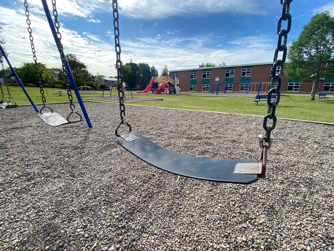 A swingset with rubber seats held up by metal chains is shown in the foreground with a long, low brick school building in the background. 
