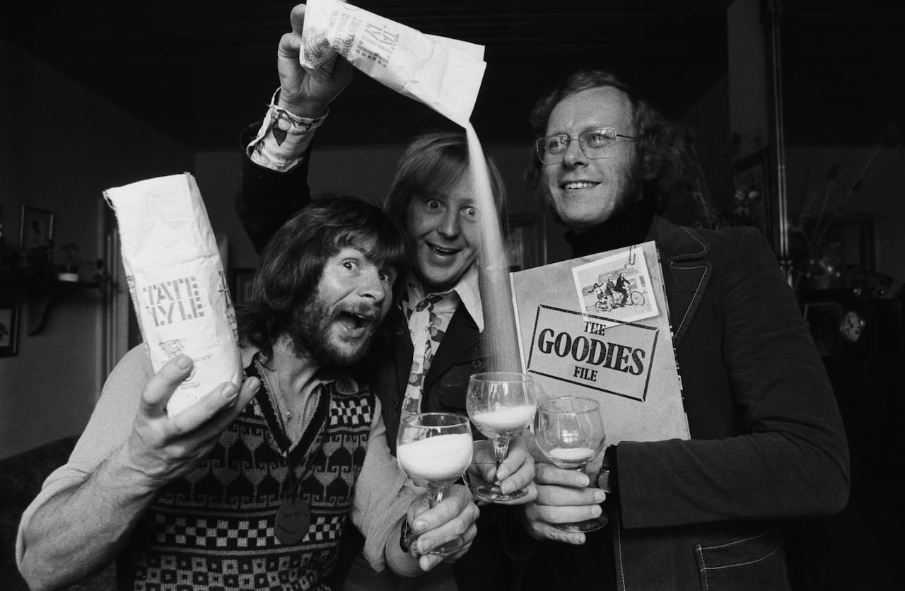 Three men are shown in a black and white photo that appears decades old. They are hamming it up in the picture, holding objects and pouring drinks.