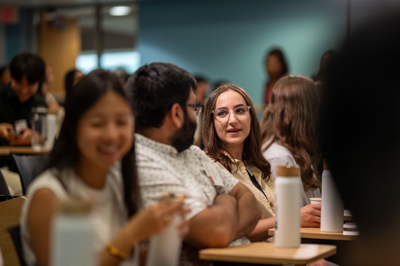New SFU medical students socialize before class starts on Wednesday. A woman with brown hair at a desk is in focus.