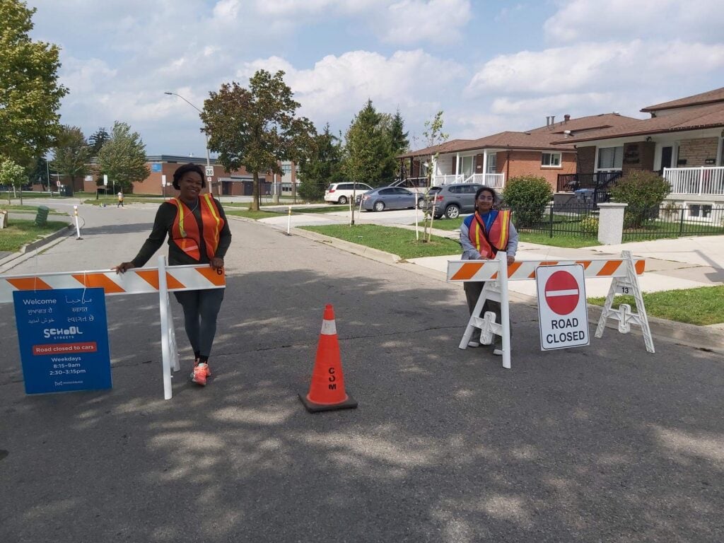 Two women stand behind temporary barriers and a sign that indicates the road is closed. They are wearing high visibility vests.