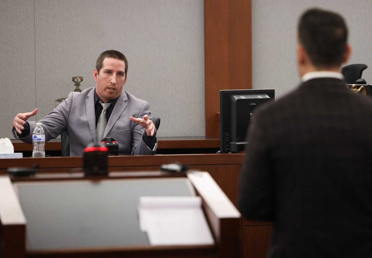A man in a suit speaks and gestures in a witness box in a court room
