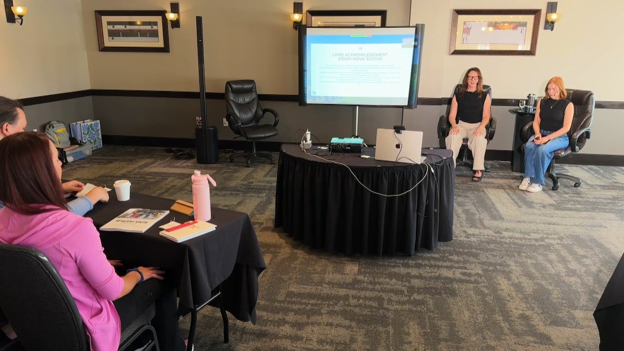 Jenny and Sophia sit at the front of the room facing the people in attendance with a presentation on a projection screen beside them.