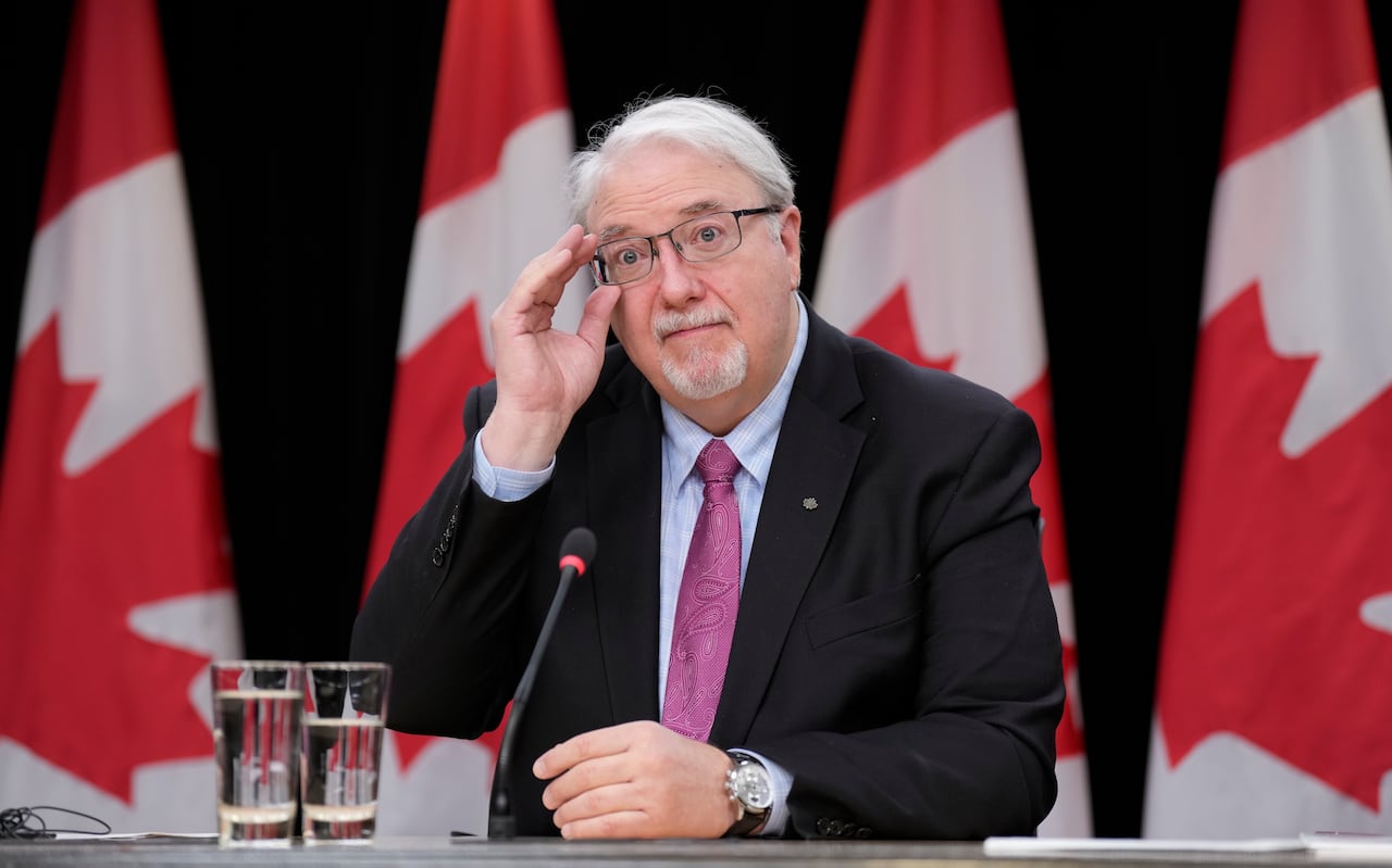 A man with white hair adjusts his glasses while wearing a suit and sitting at a table in front of four Canadian flags. Two glasses of water sit on the table next to him and a microphone is angled toward his face.