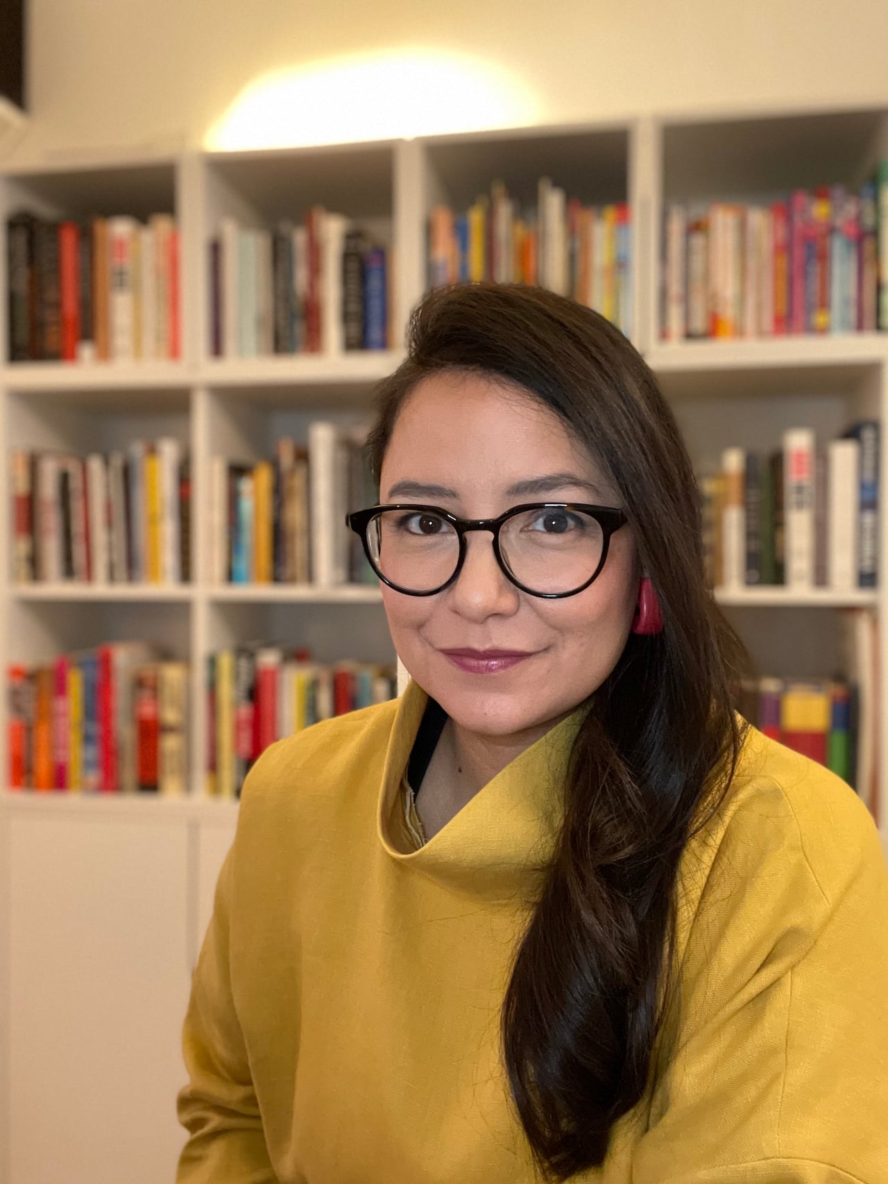 一位戴眼镜的原住民女性，背景是满载书籍的书架，她穿着芥末黄高领毛衣。