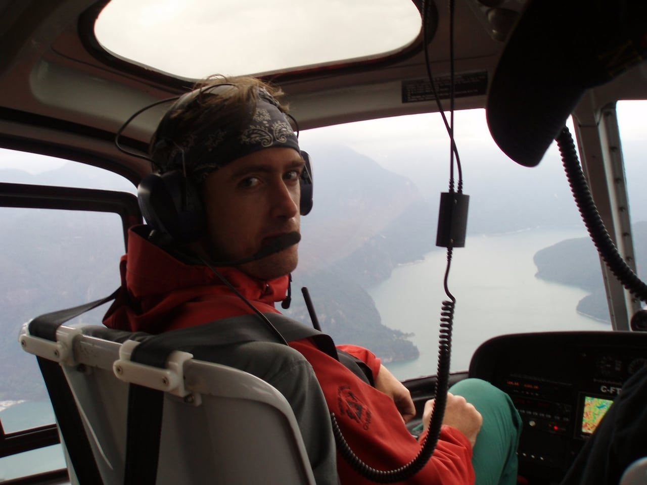 A man in a dark-coloured bandana and orange jacket is pictured in a helicopter, looking back at the camera.