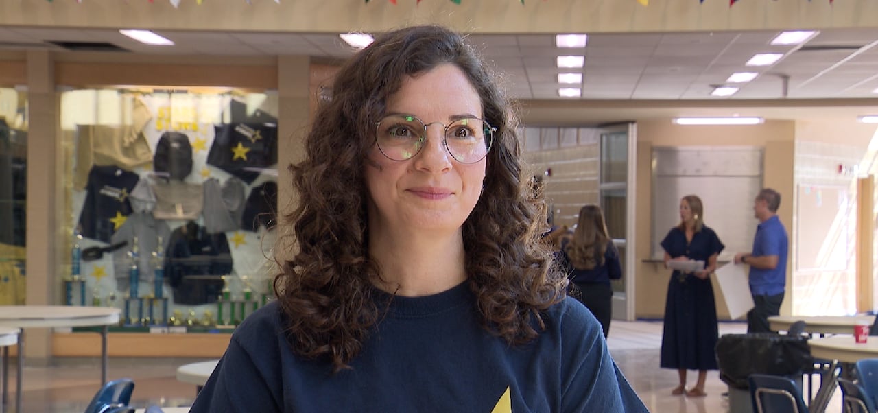 A woman with curly hair and glasses looks forward with a content expression in a school common area.