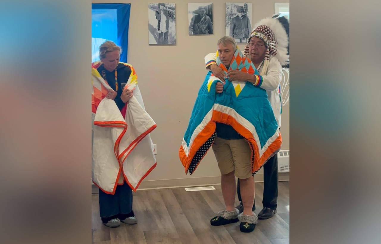 Two people are adorned in First Nations blankets by a chief in a traditional headdress