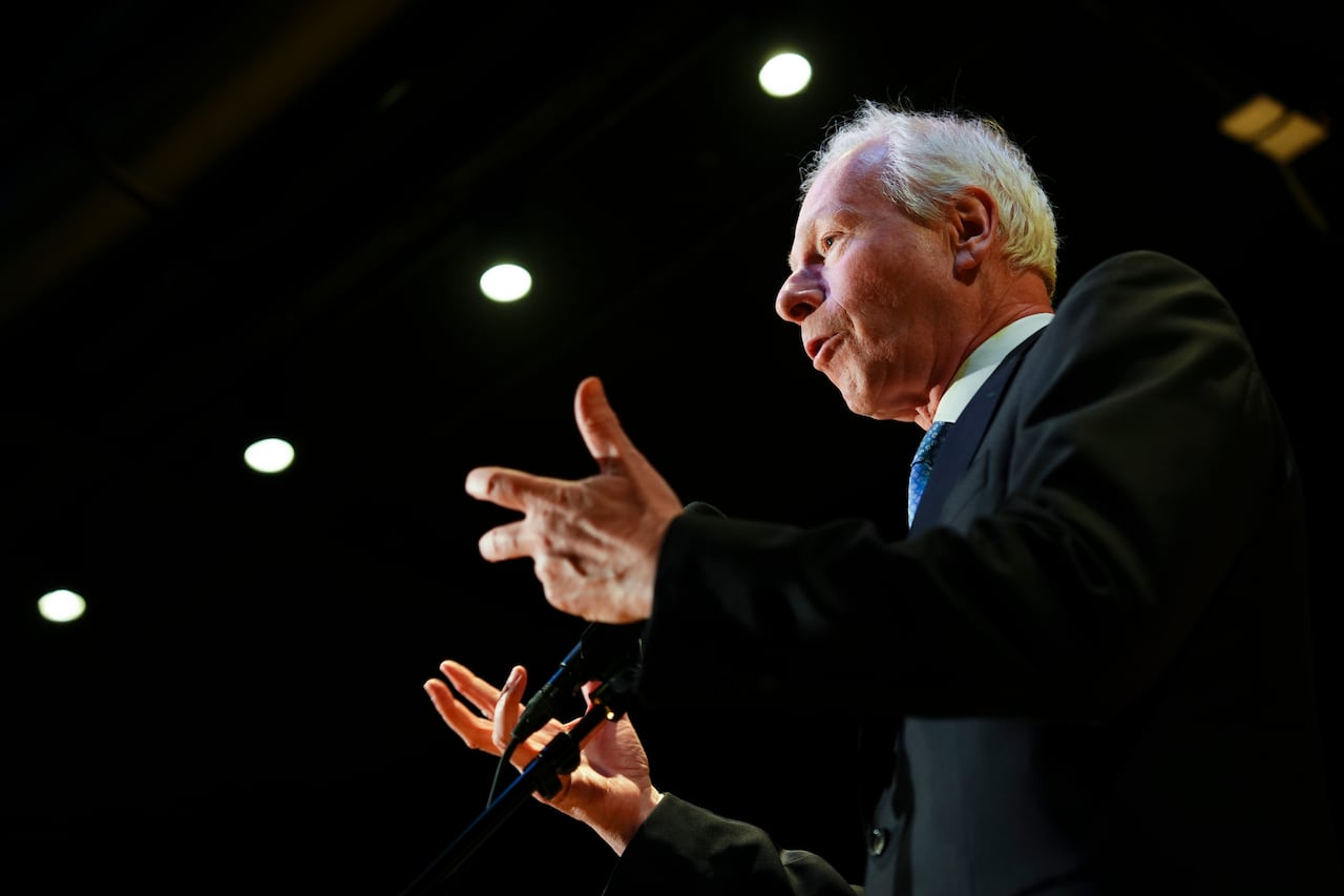 A man with short white hair wearing a dark suit is seen against a dark background with circular overhead lights seen above.