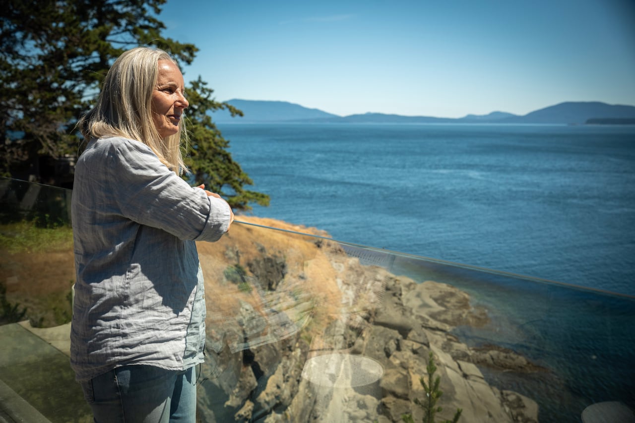 Maureen is an older white woman with blondish white hair that goes past her shoulders. She's in a light blue linen shirt. Her arms are crossed resting on a glass ledge as she look out to the Salish Sea.