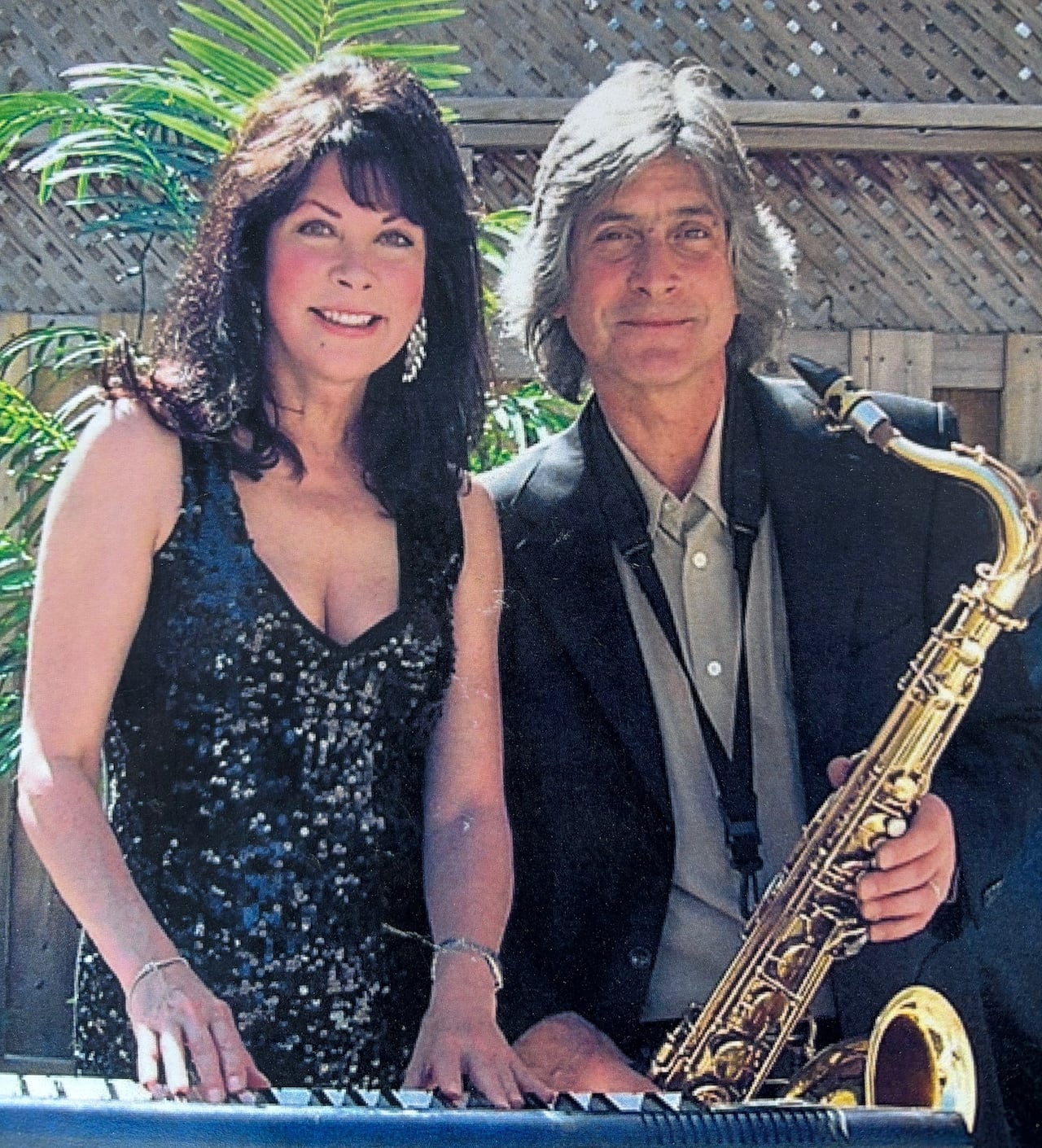 A woman in a sparkly black dress and a man holding a saxophone stand behind a keyboard in front of a palm leaf.