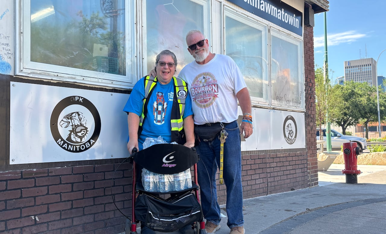 A woman with a walker wears a bright yellow west vest. A man in a white t-shirt stands next to her