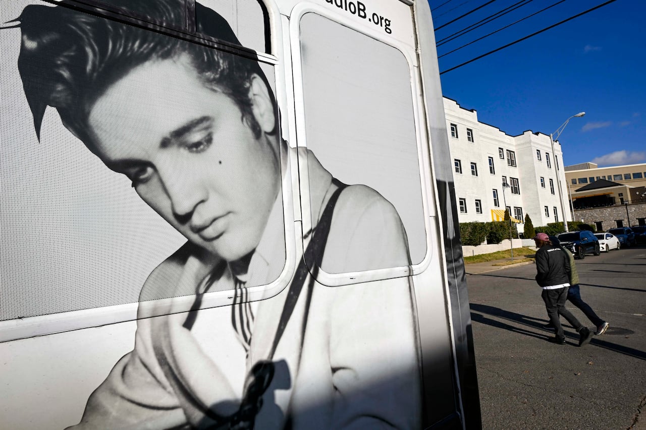 An image of a young man with dark hair looking down as he plays guitar appears on the side of a bus, with two people crossing the road behind it.