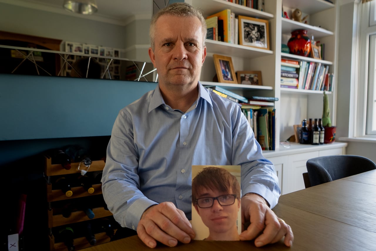 A man with short silver hair sits at a table in front of a bookshelf holding a photo of a young man with short hair and glasses.