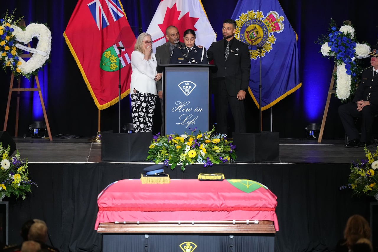 Woman speaks with three people behind her at podium with casket on the ground below.