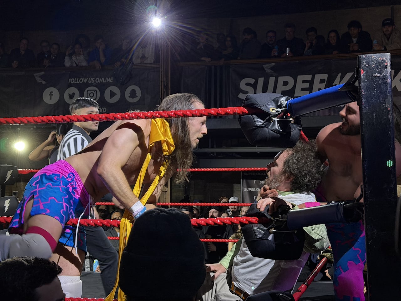 Two wrestling opponents face off in the ring, one of them appears to be knocked out and laying on the ropes.