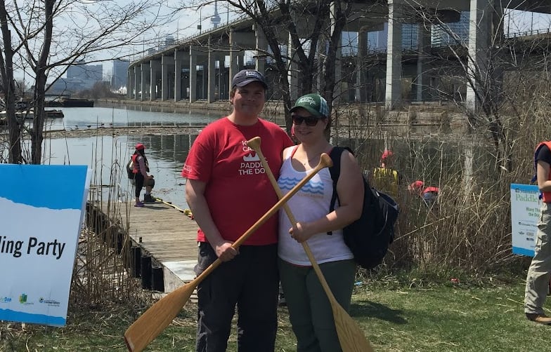 A man and woman stand with canoe paddles, open water and the Toronto skyline is in the background.