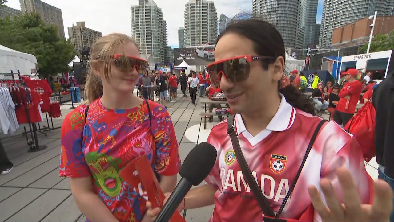 A young man and woman in sunglasses and Canada soccer jerseys talk into a reporter's microphone at a festival in downtown Toronto on a cloudy spring day
