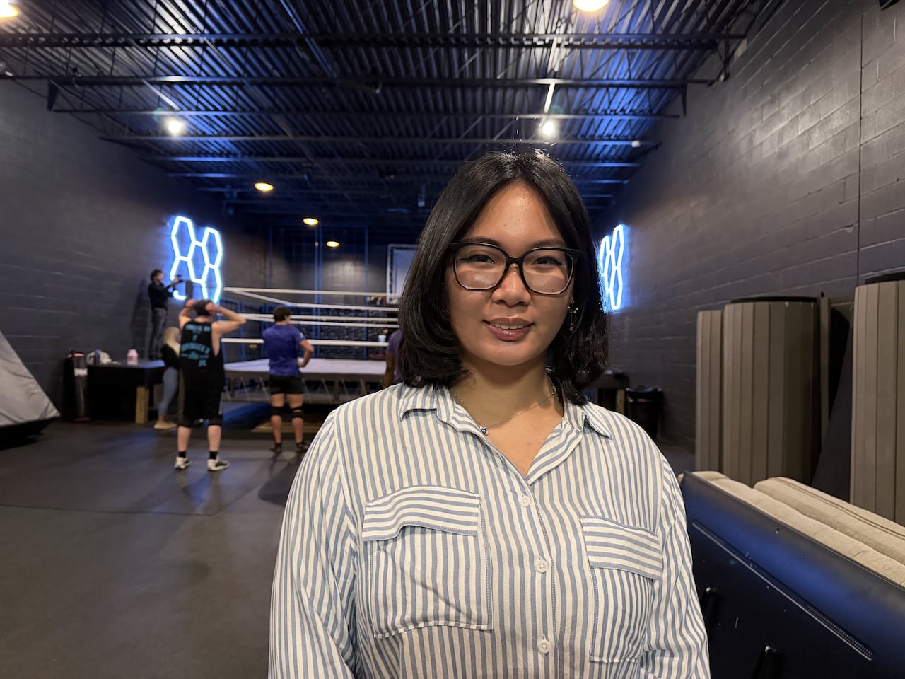 A woman with short black hair and glasses standing in a wrestling gym, smiling.