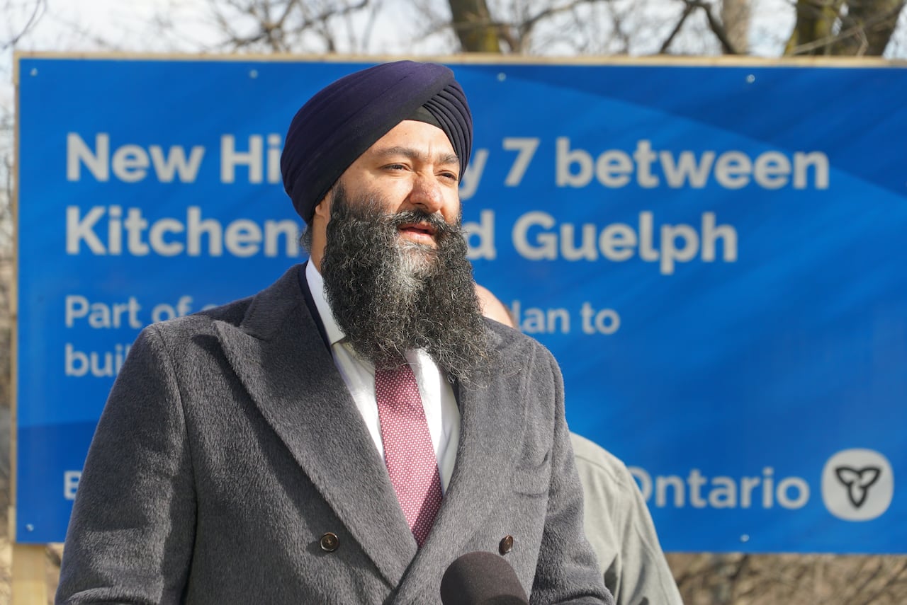 A man wearing a suit and red tie speaking in front of a blue sign.