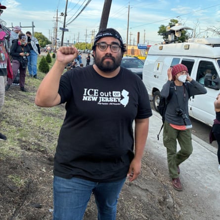 a man in a black ‘ICE out of new jersey’ T-shirt holds up a fist while standing outside at protest